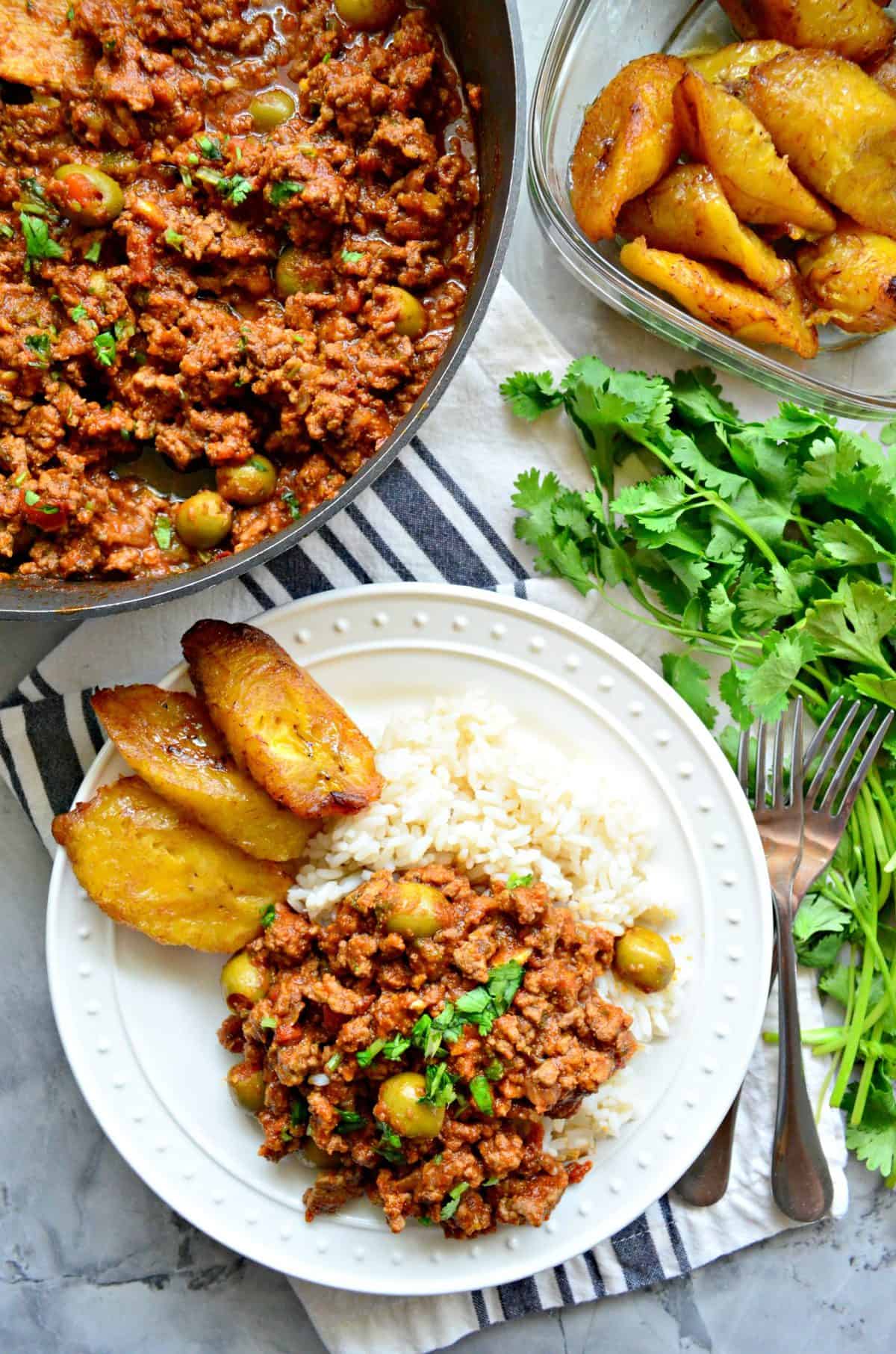 top view plated ground meat, red sauce, green olives, and fresh herbs served with rice and plantains.