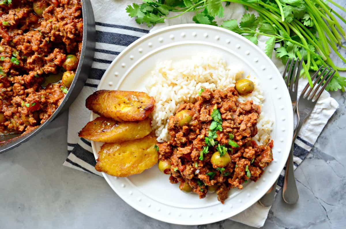 top view of ground meat, red sauce, green olives, and fresh herbs served with rice and plantains on plate.