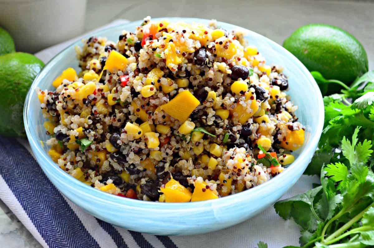 quinoa with corn, peppers, cilantro, and mango in blue bowl next to fresh cilantro and limes.