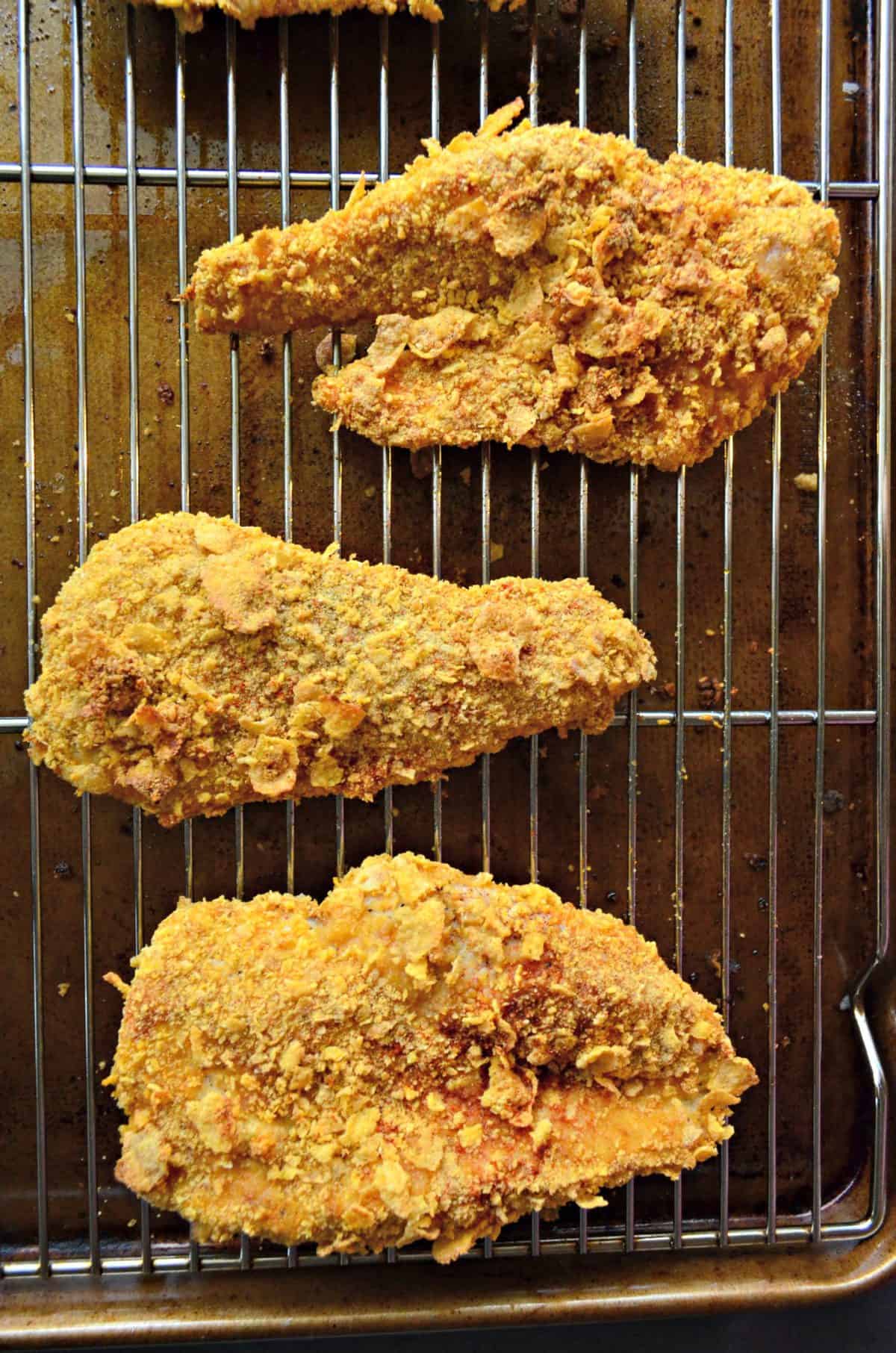 top view 3 chicken thighs or breasts coated in corn flakes on rack over top of sheet pan.