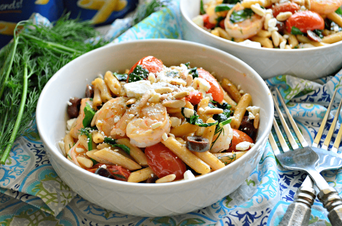 close up bowl of penne with pine nuts, spinach, shrimp, tomatoes, kalamata, and cheese visible with forks.