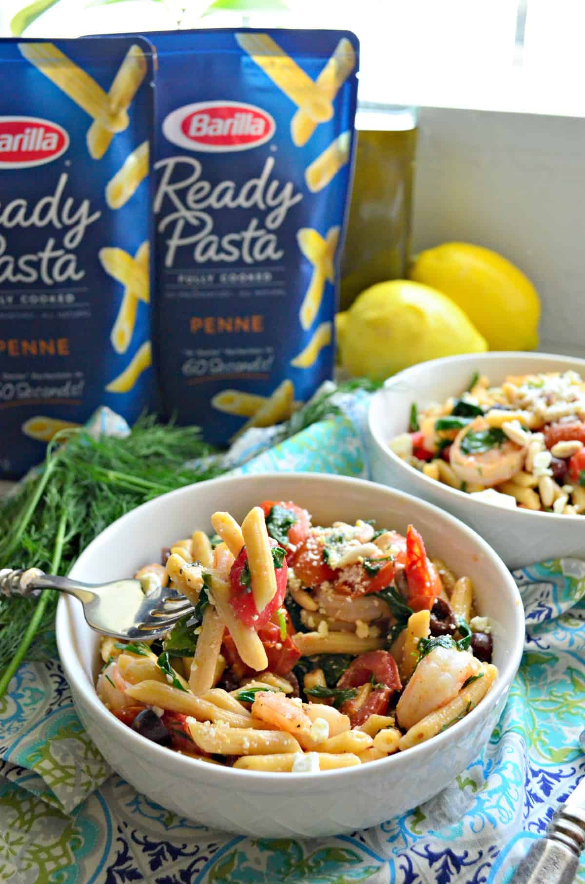 two bowls of penne with pine nuts, spinach, shrimp, tomatoes, kalamata, and cheese by ready pasta packages.