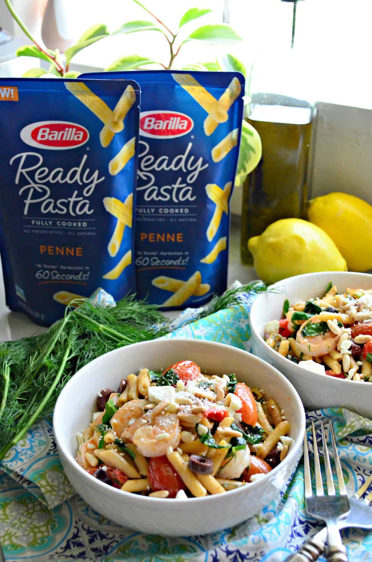 two bowls of penne with pine nuts, spinach, shrimp, tomatoes, kalamata, and cheese on placemat with fresh dill.
