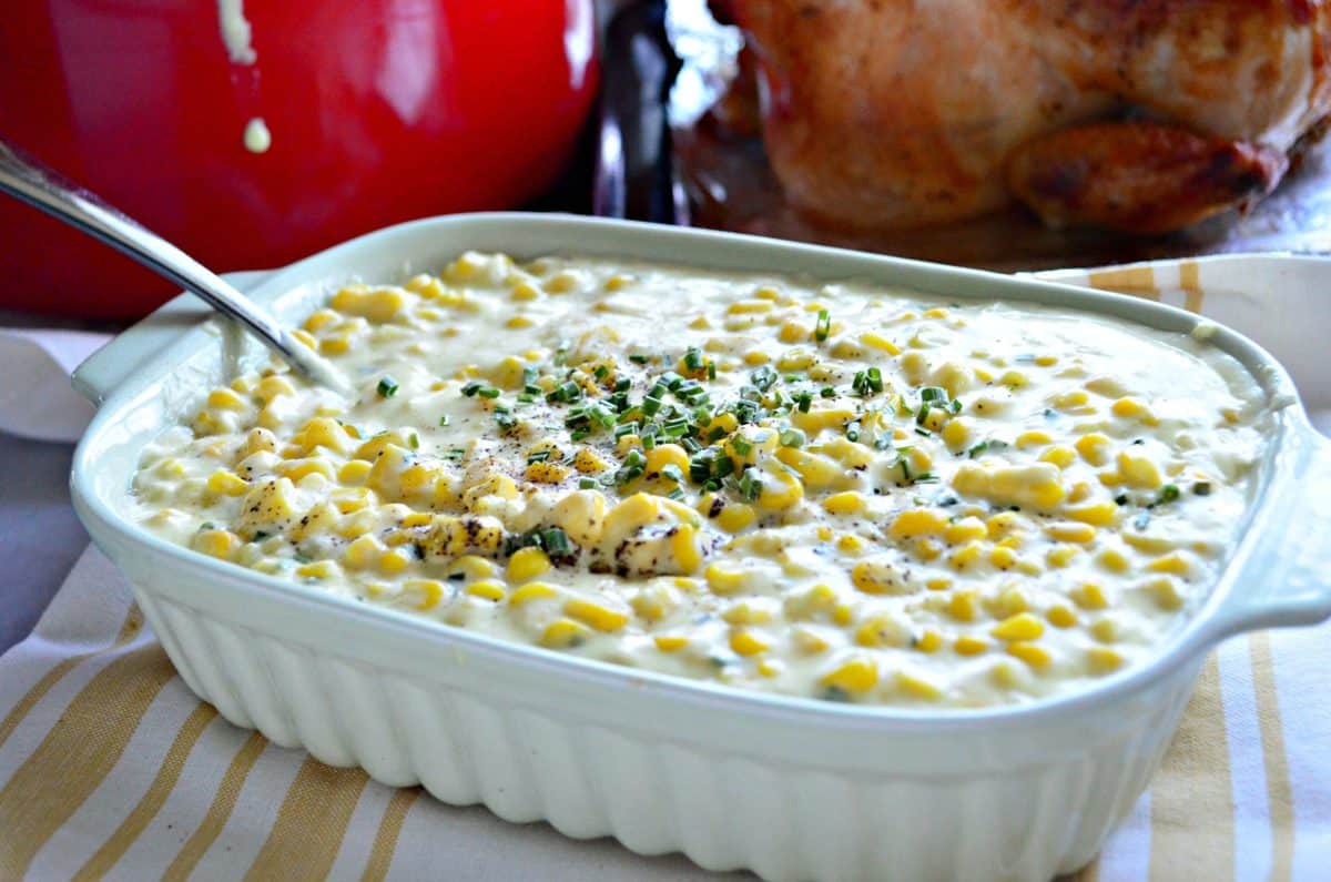 creamy corn with pepper and herbs in a casserole dish with a silver spoon on a yellow striped tablecloth.
