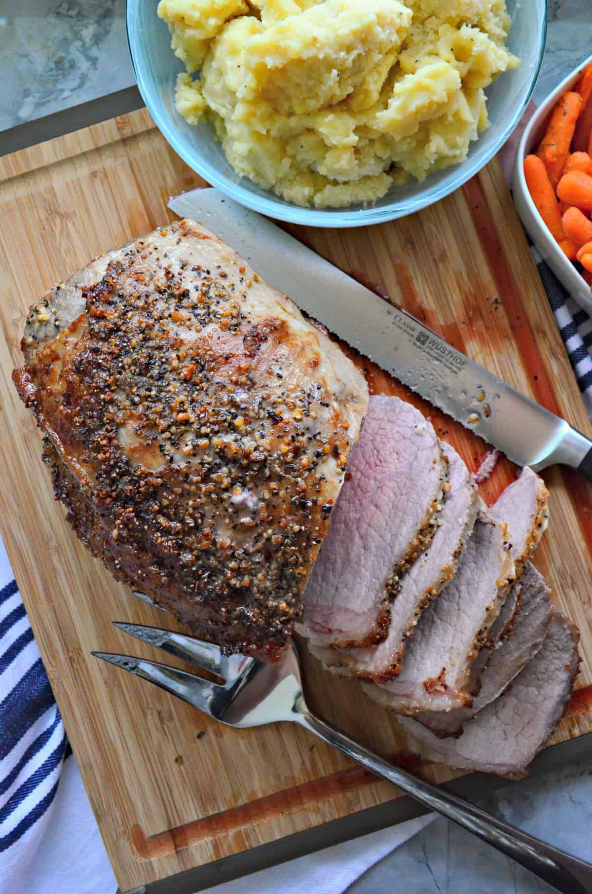 top view sliced roast beef crusted with spices on cutting board with bowl of mashed potatoes.