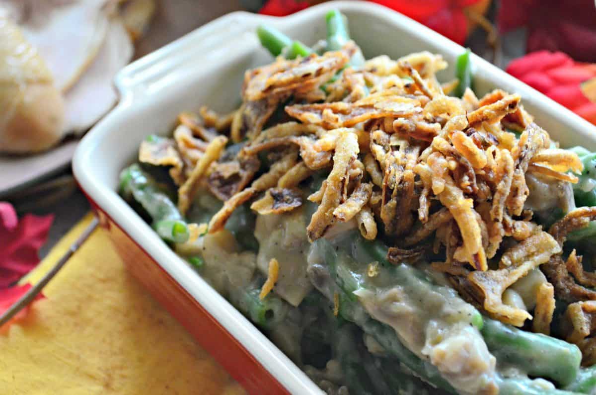 side view of corner of small casserole dish of green beans with white chunky substance topped with crispy onions. 