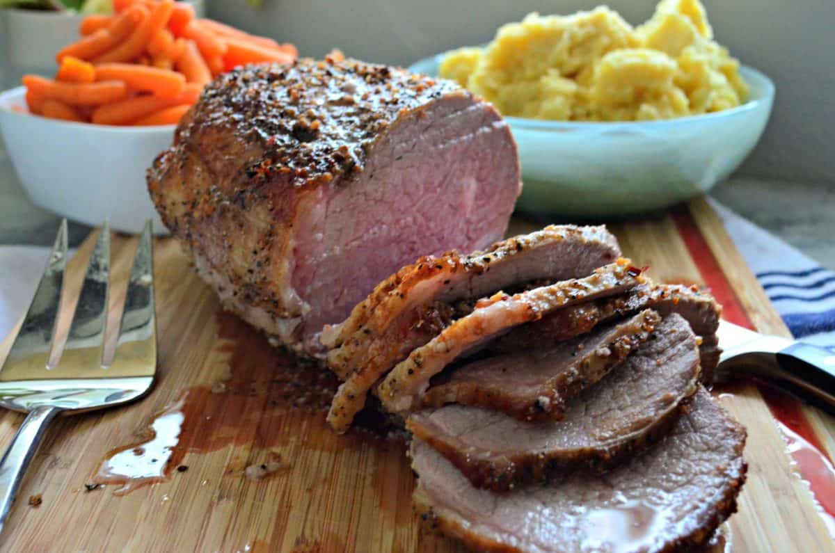 sliced roast beef crusted with spices on cutting board in front of carrots and mashed potatoes next to fork.