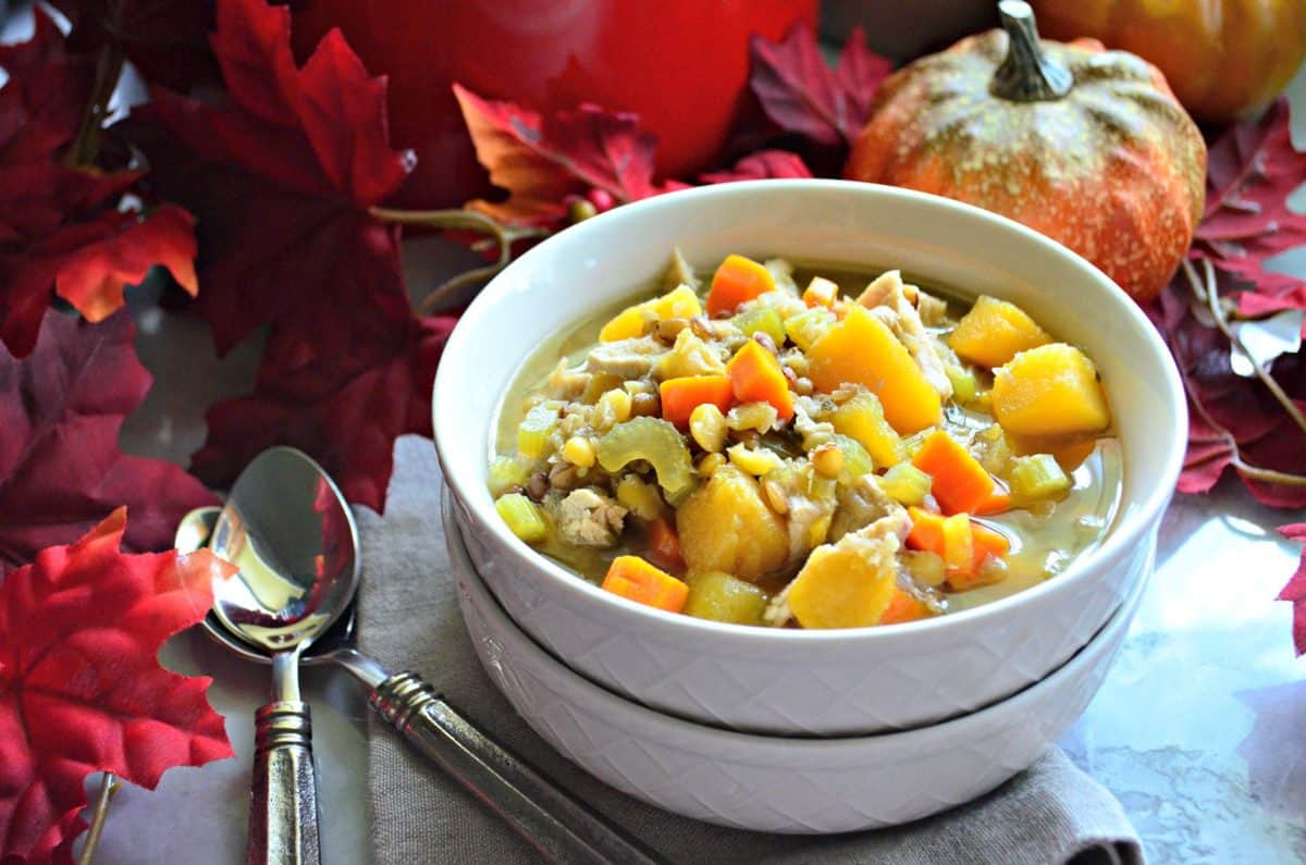 Side view of Turkey Butternut Squash and Lentil Soup in white bowl on table with fake decorative fall leaves and pumpkins.