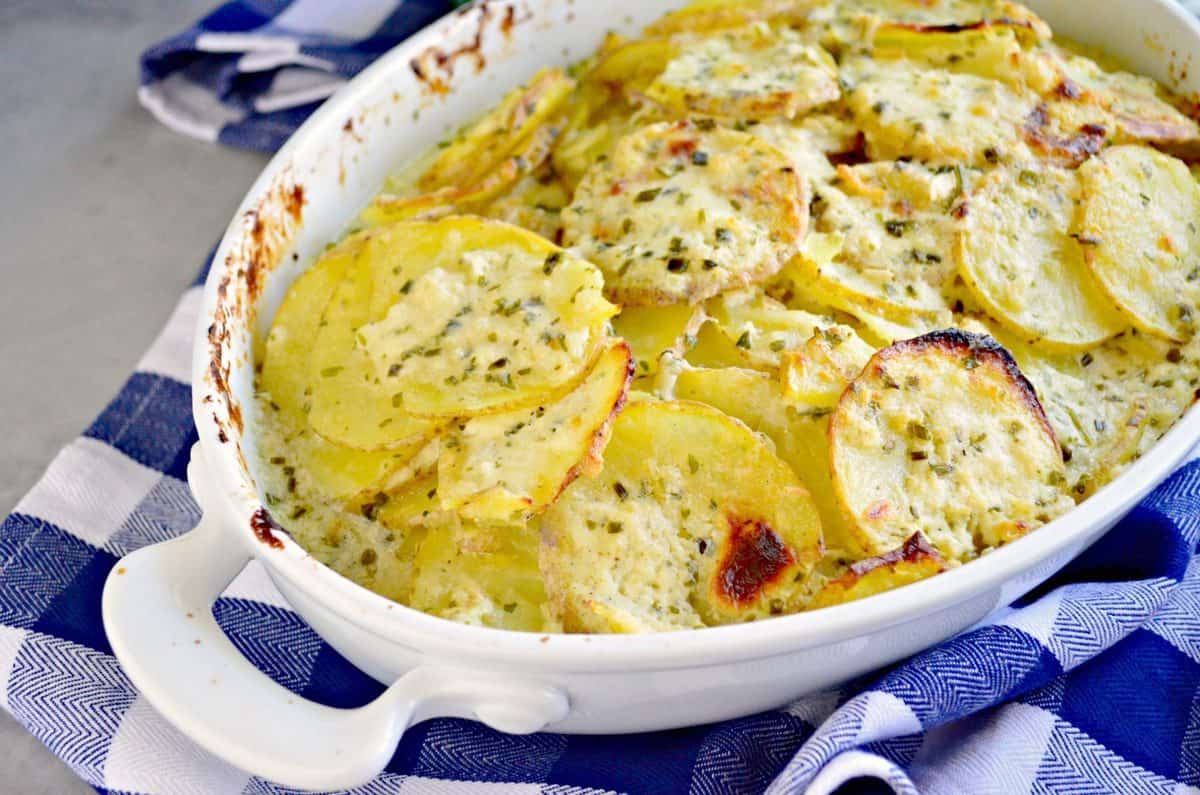 creamy and golden brown scalloped potatoes with herbs and cheese in white ceramic dish.