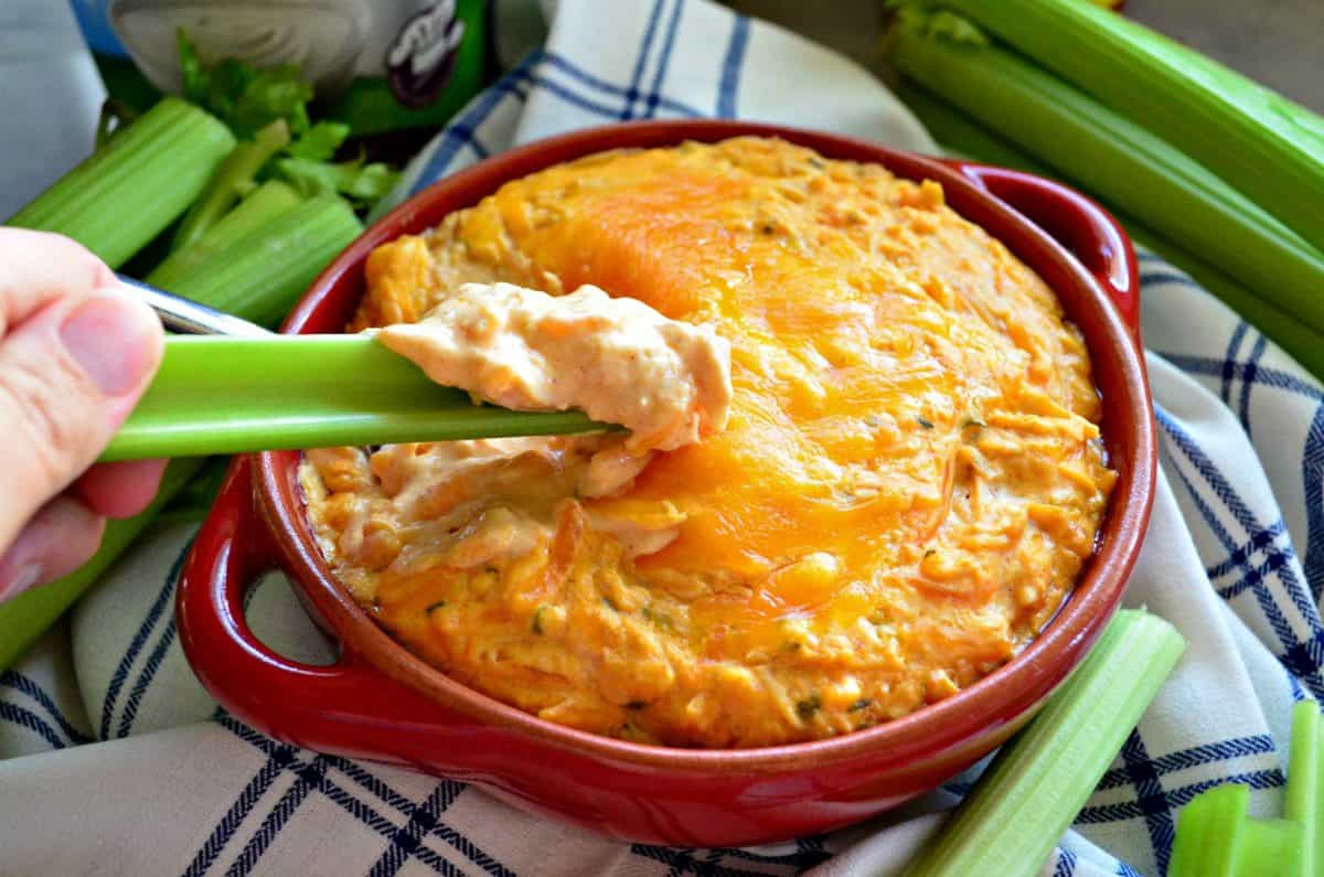 hand dipping piece of celery into thick orange cheesy dip in red bowl.