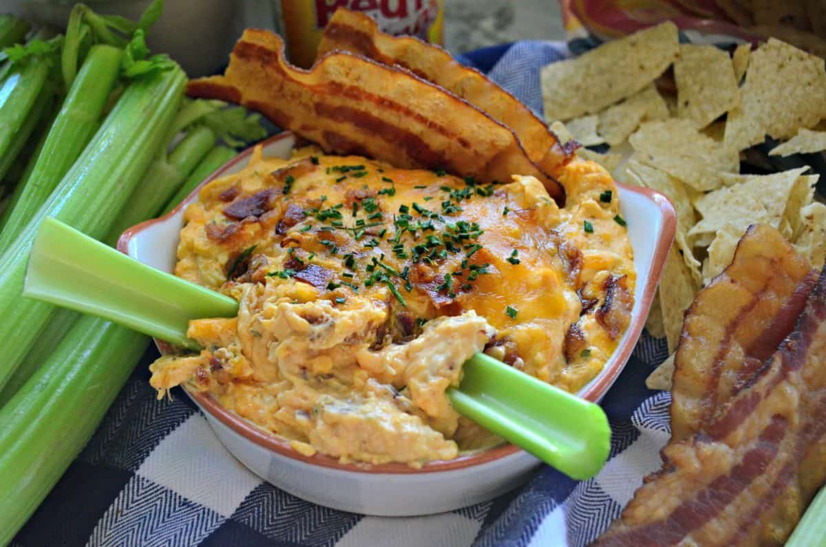 bowl of thick orange colored dip with bacon bits and 2 celery sticks sticking out.