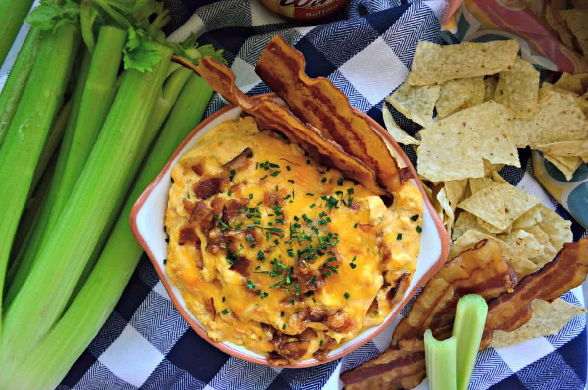 top view bowl of thick orange colored dip with bacon bits and herbs next to chips and celery.