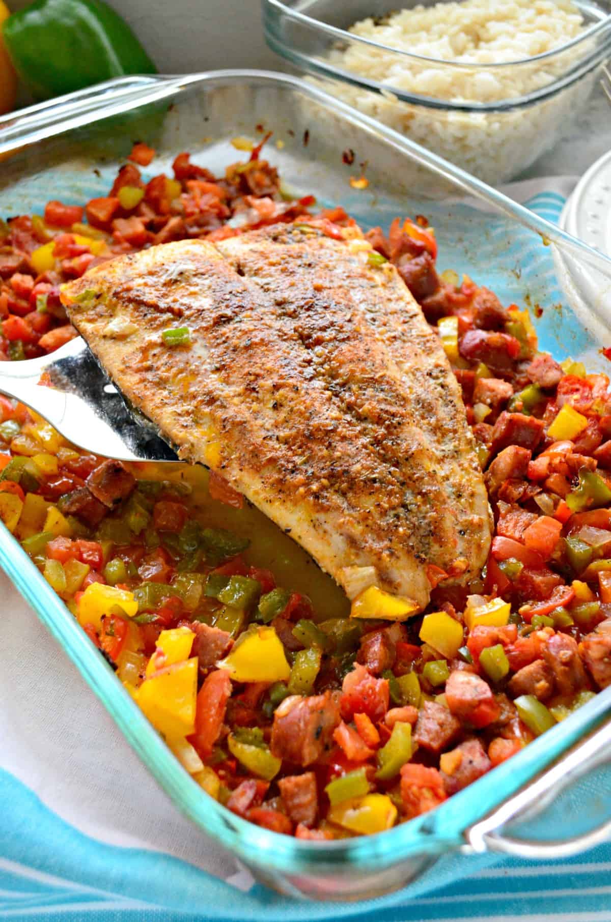 whole baked mahi mahi held up with spatula over chopped veggies and sausage in glass baking dish.