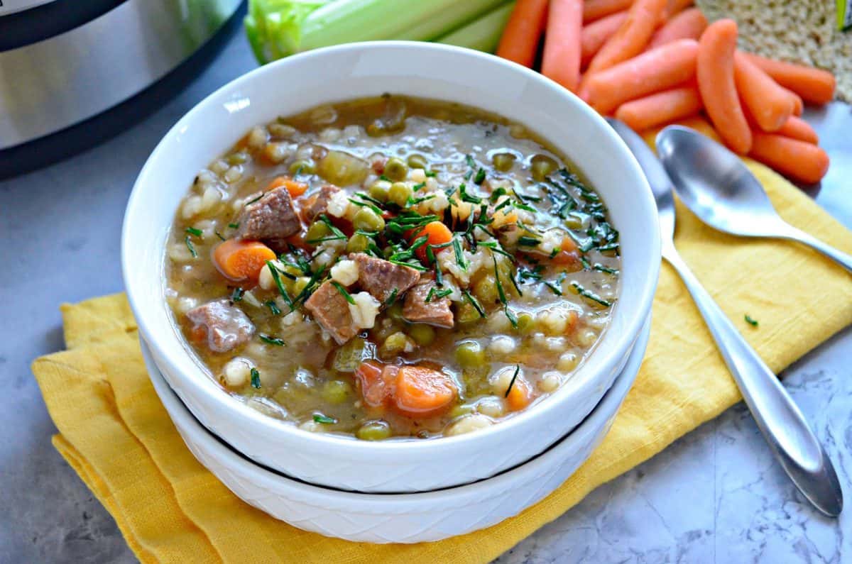 top view of bowl of brown soup with barley, beef, peas, carrots, celery and herbs visible next to spoons.