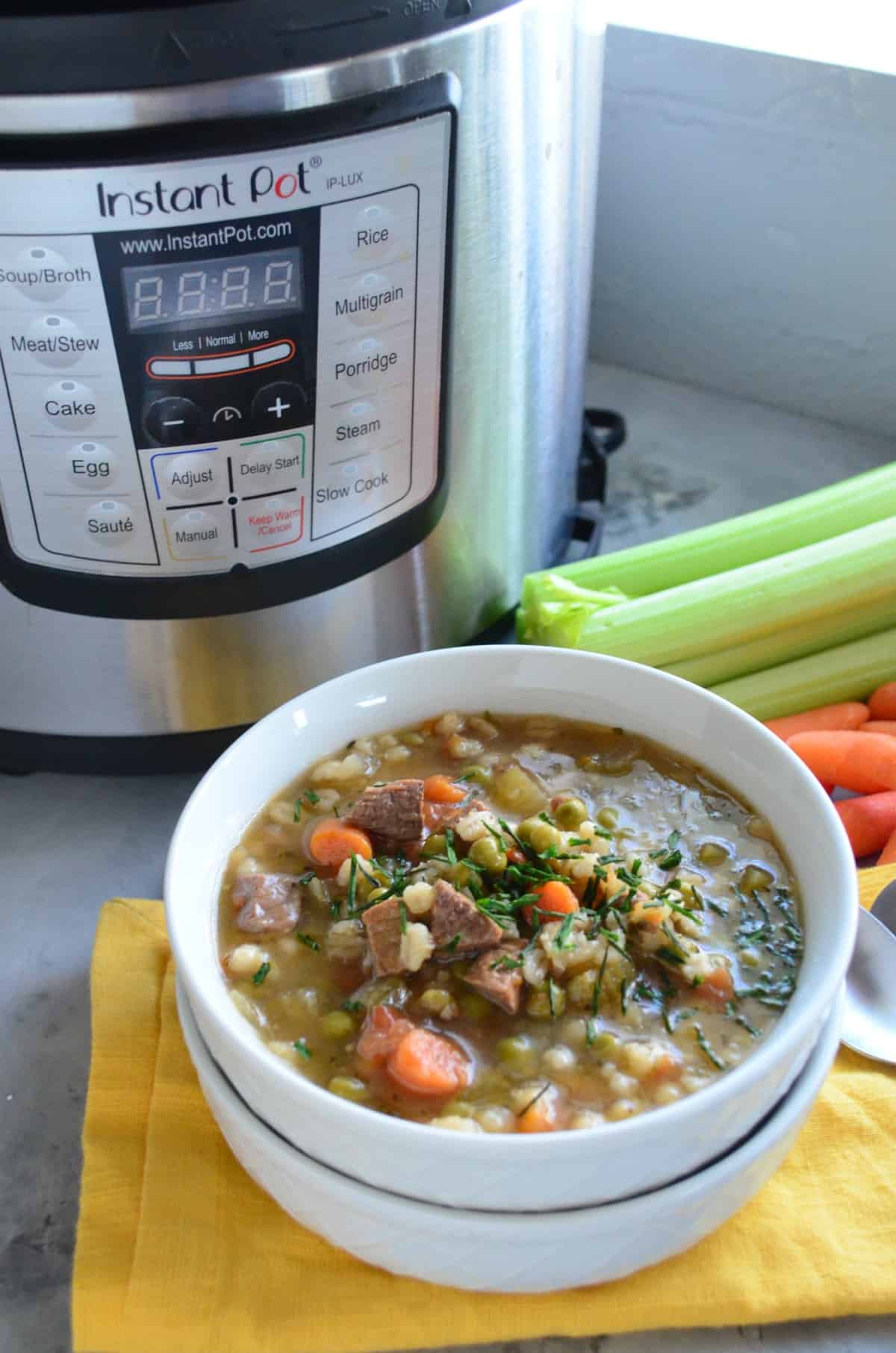 top view of bowl of brown soup with barley, beef, peas, carrots, celery and herbs in front of instant pot.
