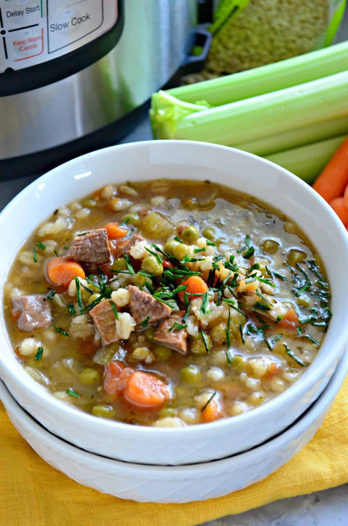 closeup bowl of brown soup with barley, beef, peas, carrots, celery and herbs in front of fresh celery.
