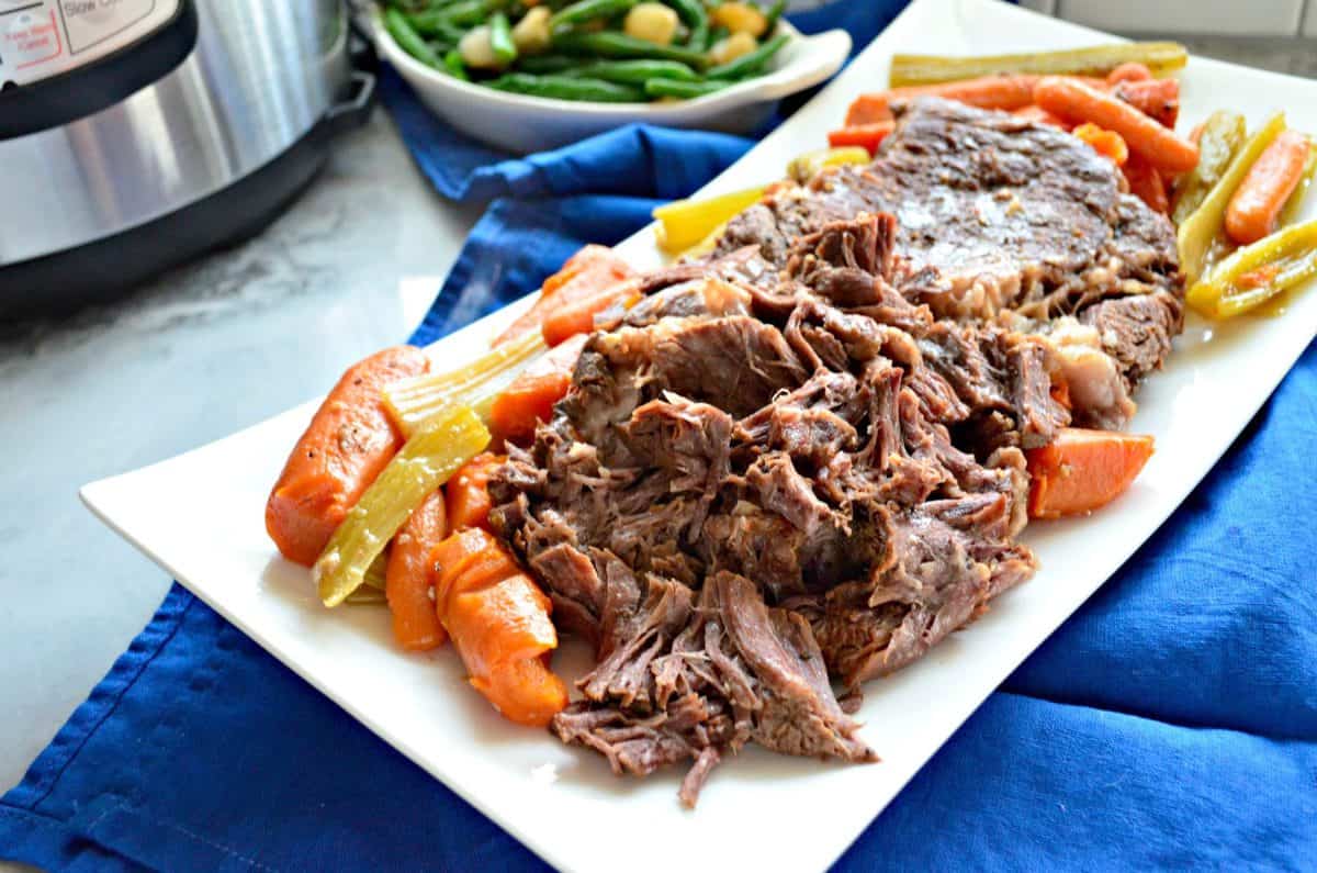 top view of rectangular platter of pot roast with cooked carrots and celery on blue tablecloth.