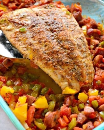 Silver spatula holding a large piece of cooked mahi over a bed of peppers and sausage.