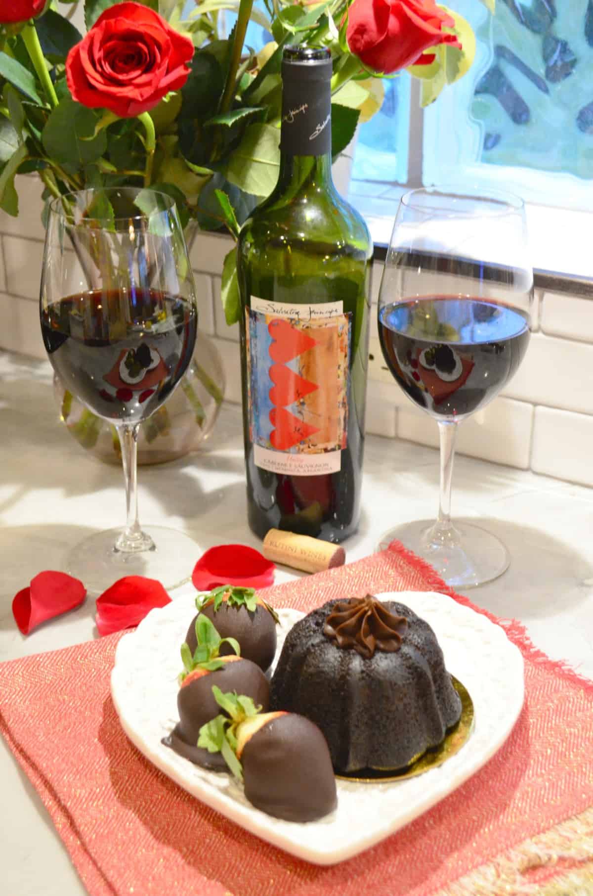 two glasses of wine and bottle behind plated chocolate cake plated with chocolate dipped strawberries.