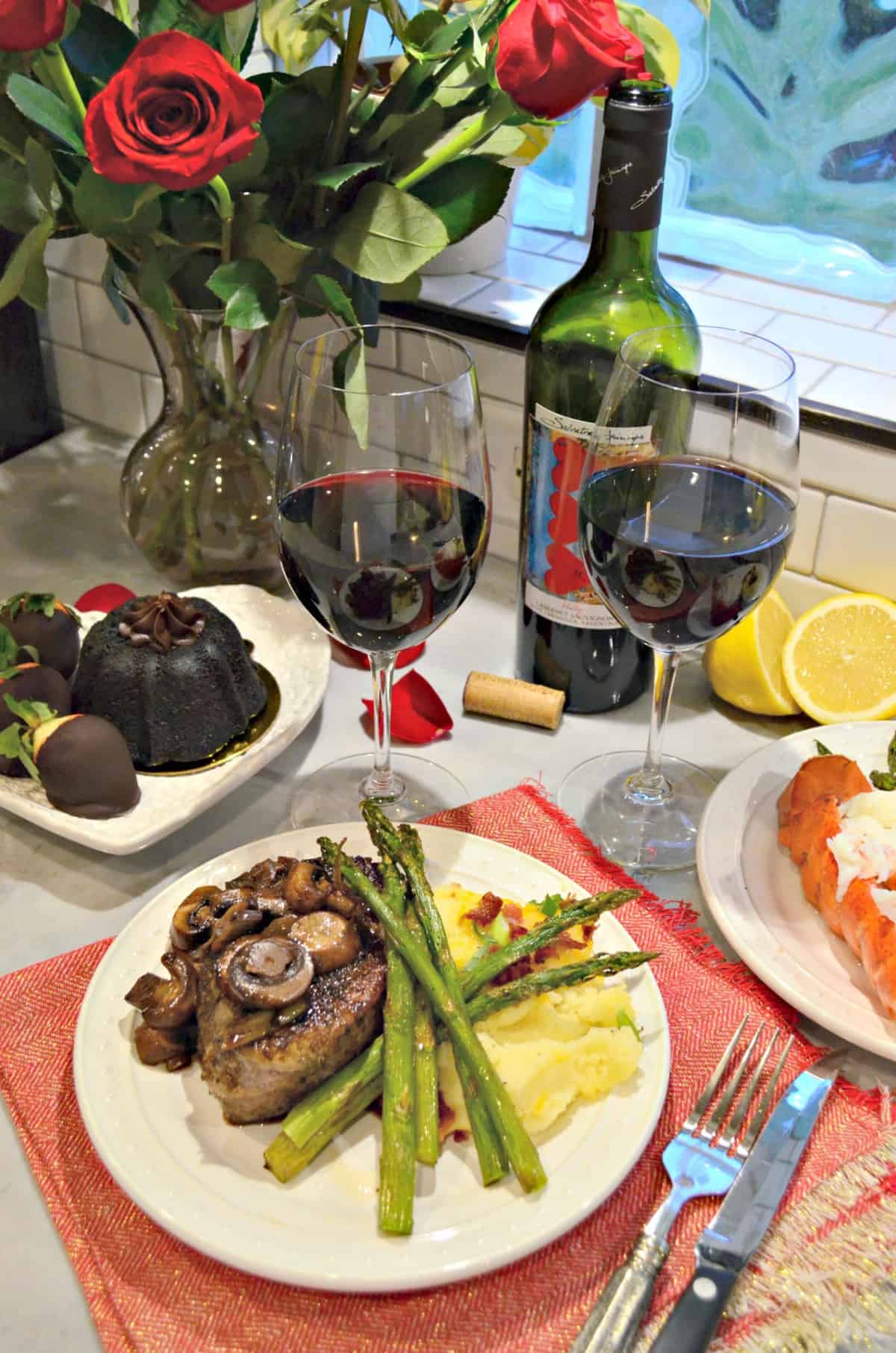 two glasses of wine next to plated chocolate cake, steak, and lobster with asparagus and potatoes.