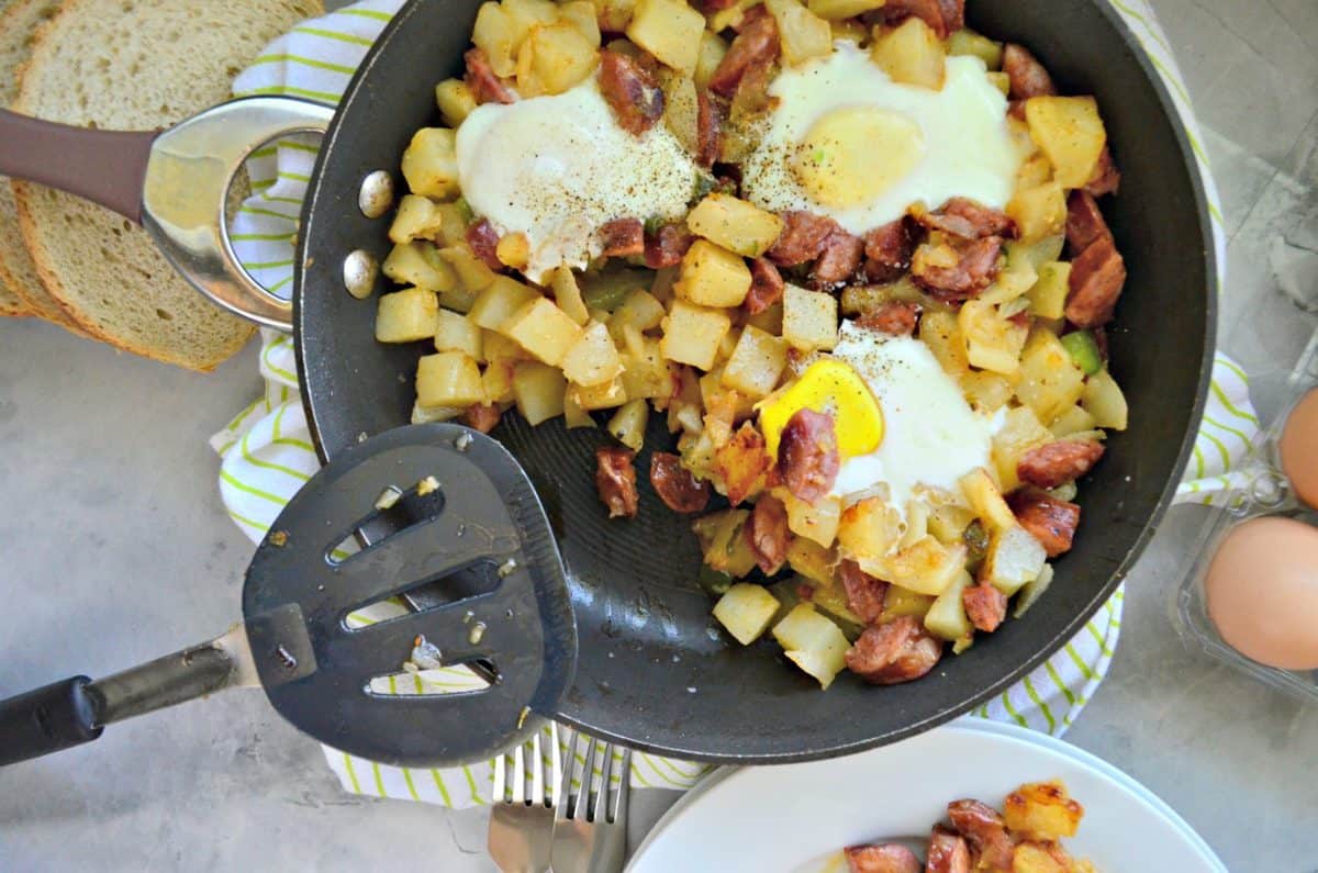 top view of diced potatoes, sausage, over easy eggs, peppers, and pepper in skillet with spatula.