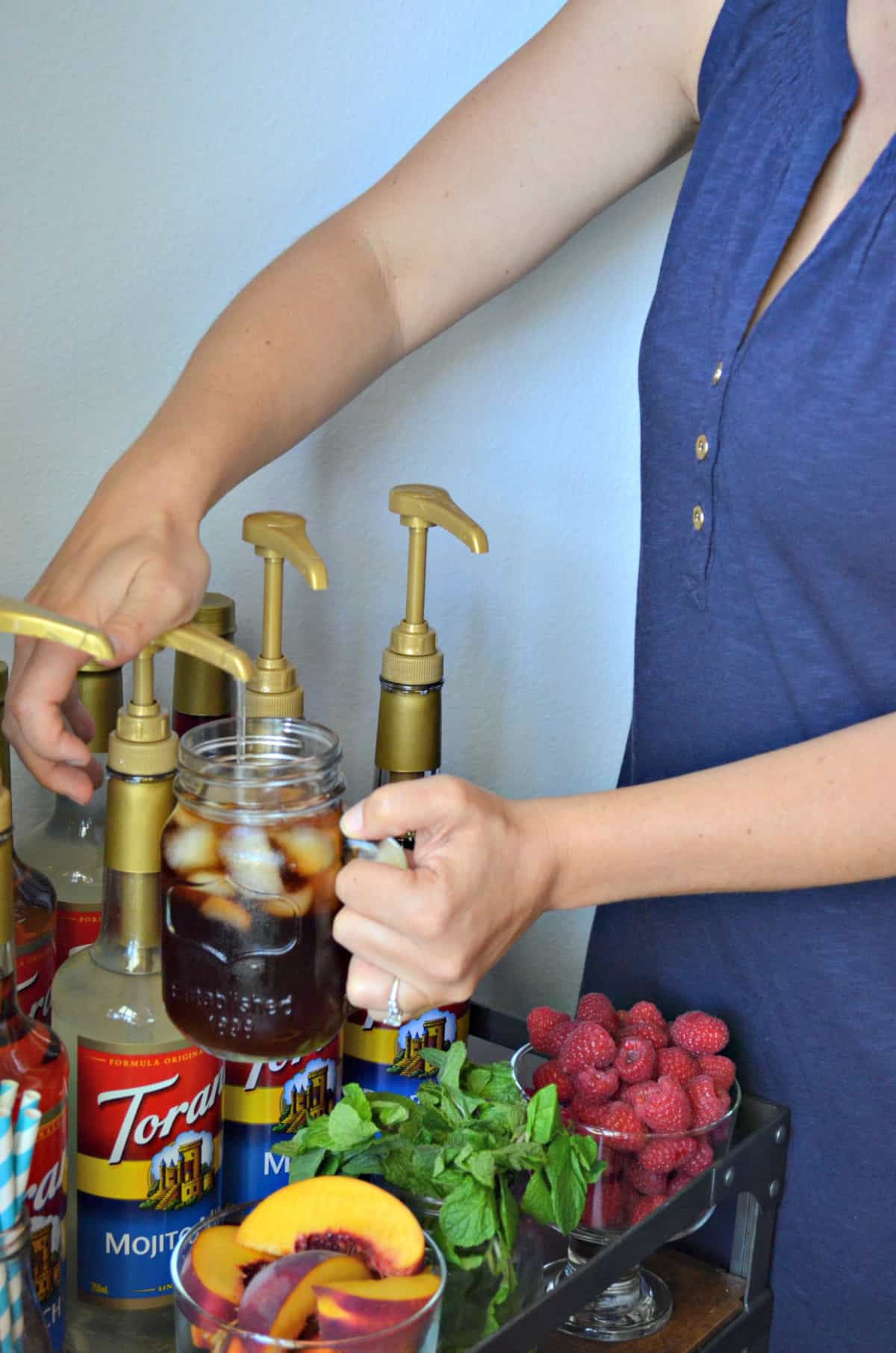 female hand pumping mojito syrup into iced tea on bar cart that has mint, strawberry, and peaches.