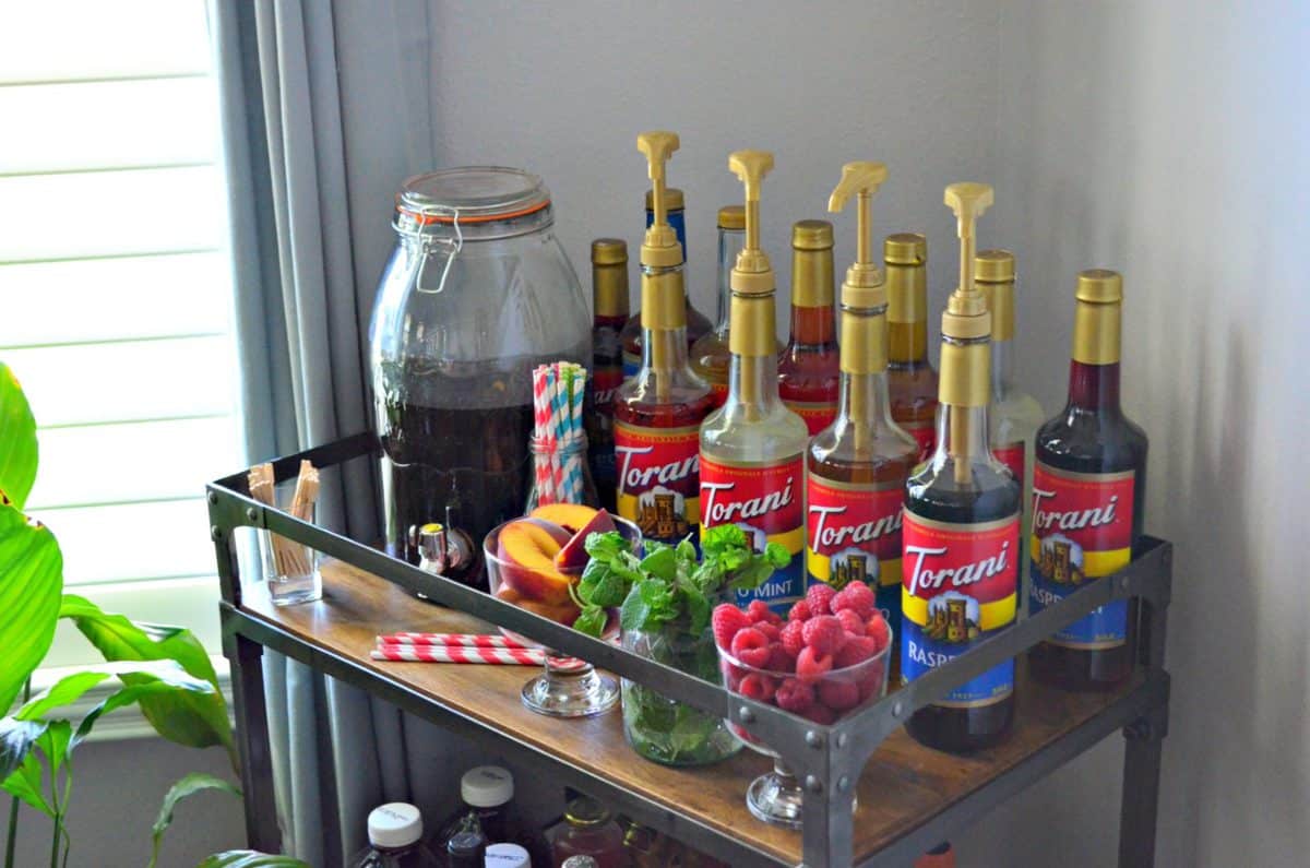 bar cart with 10 torani syrup flavors decoratively placed with mint, raspberries, peaches, and straws.