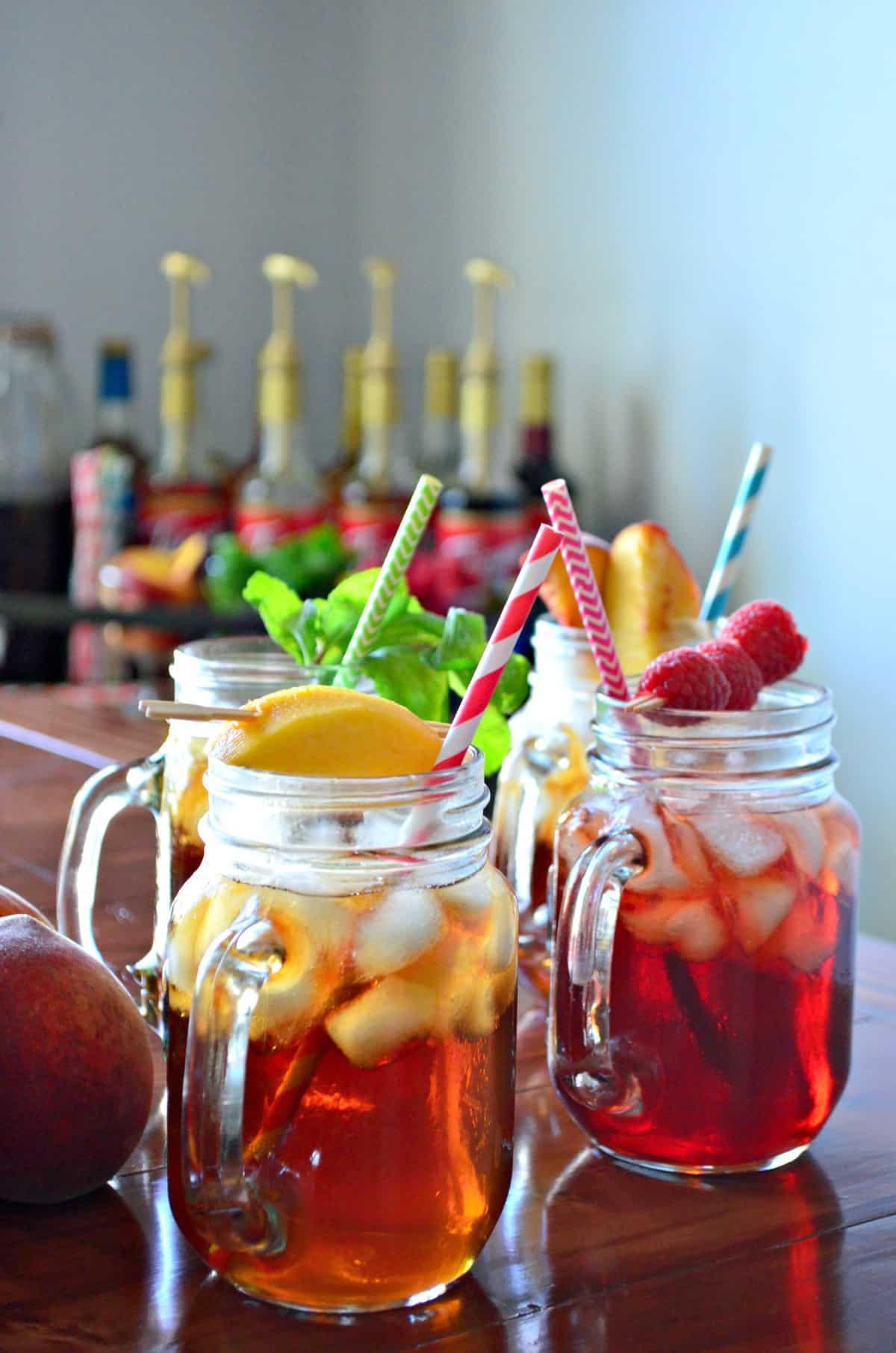 4 glass jars of iced tea, each garnished with a different item like peaches, raspberries, and mint. 