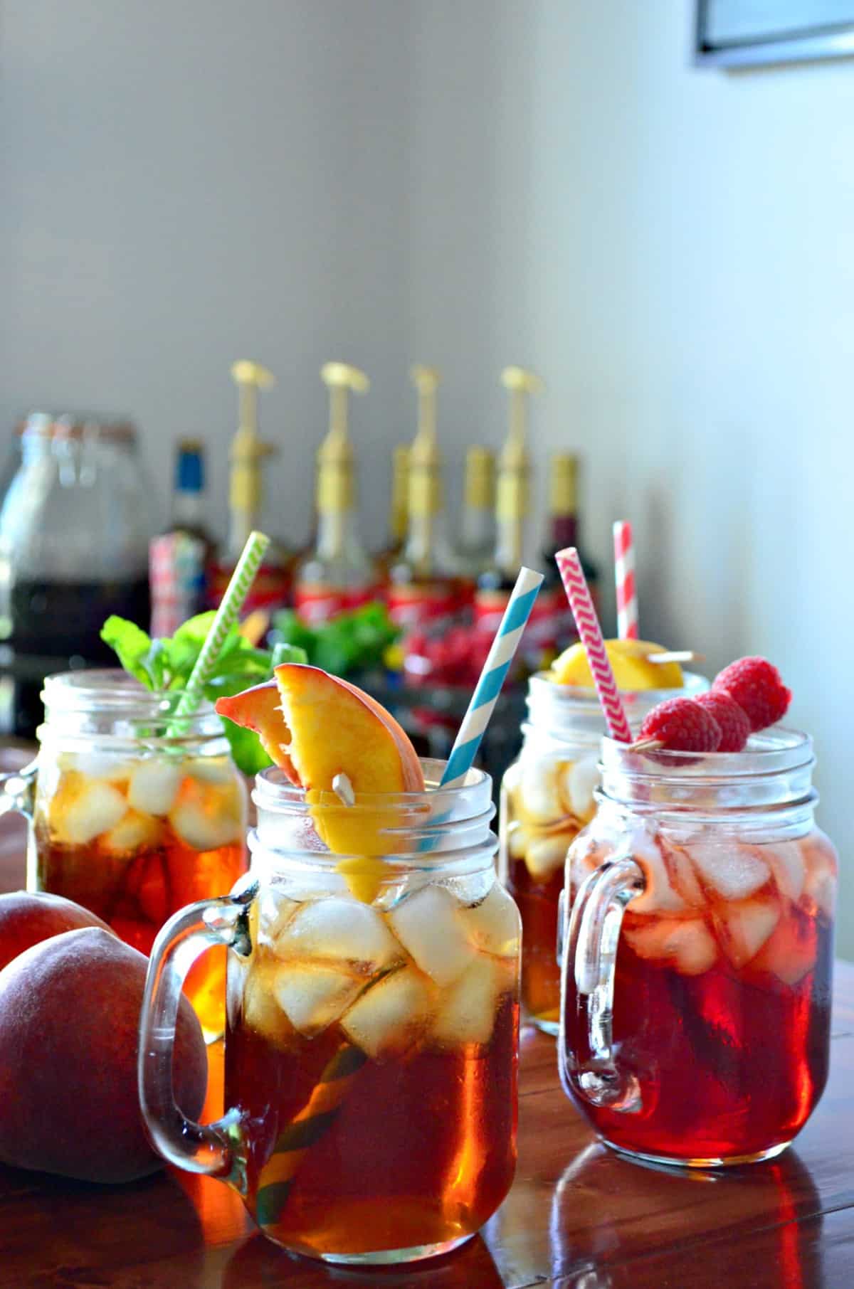 4 glass jars of iced tea, each garnished with a different item like peaches, raspberries, and mint. 