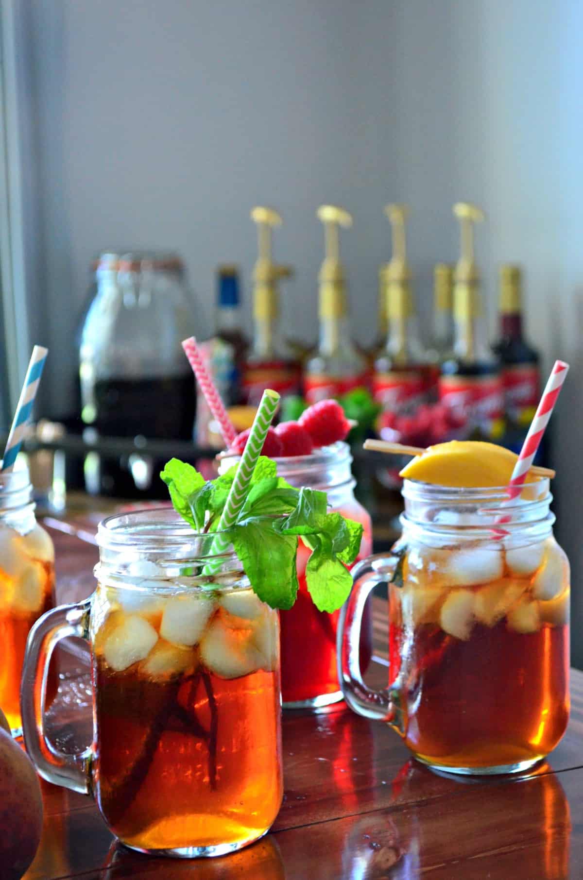 3 glass jars of iced tea, each garnished with a different item like peaches, raspberries, and mint. 