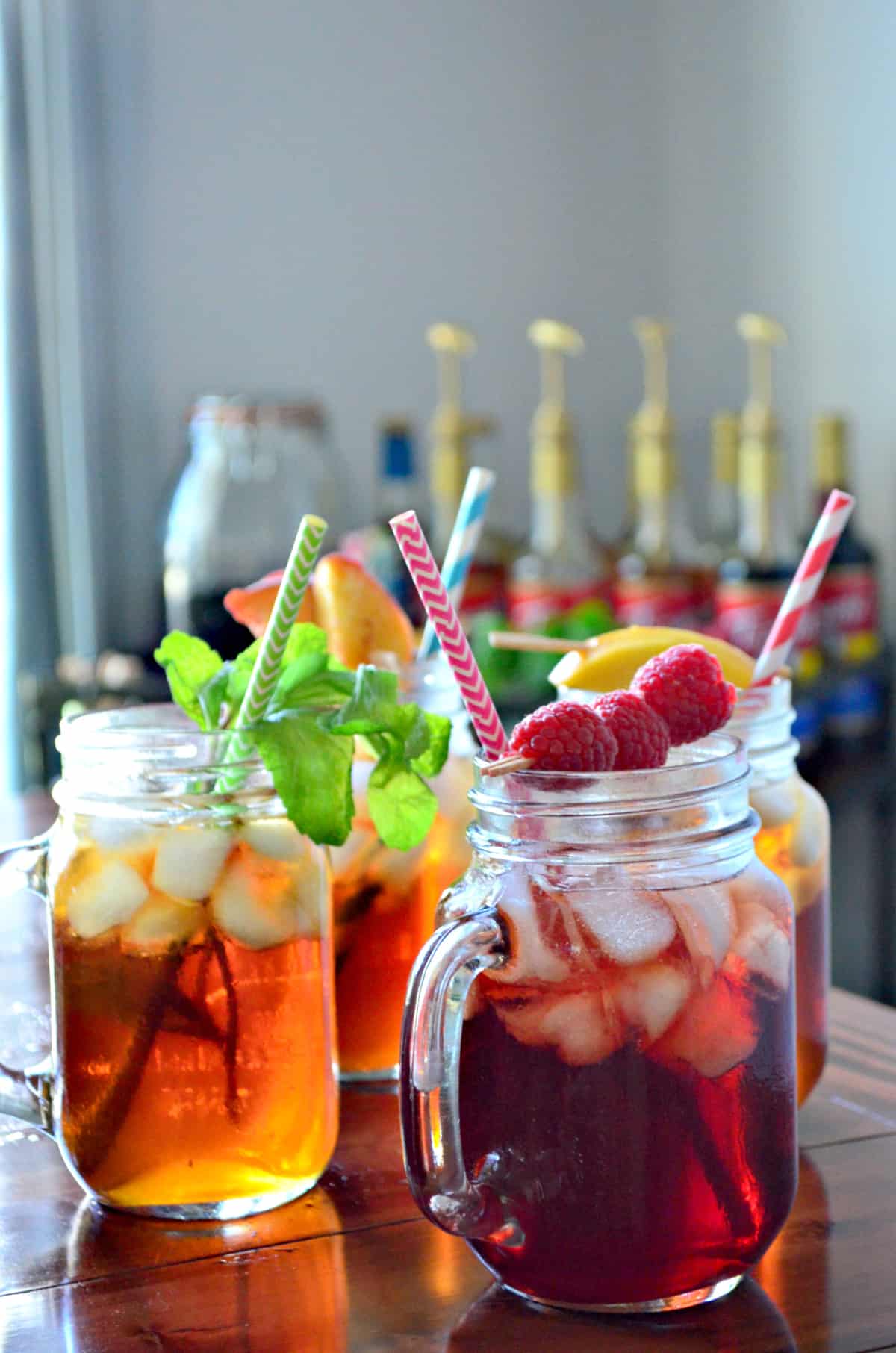 4 glass jars of iced tea, each garnished with a different item like peaches, raspberries, and mint.