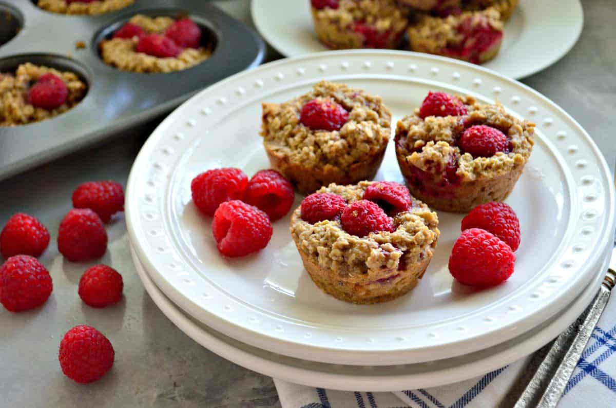 3 plated Banana Raspberry Oatmeal Muffins with raspberries in front of muffin tin and raspberries on countertop.