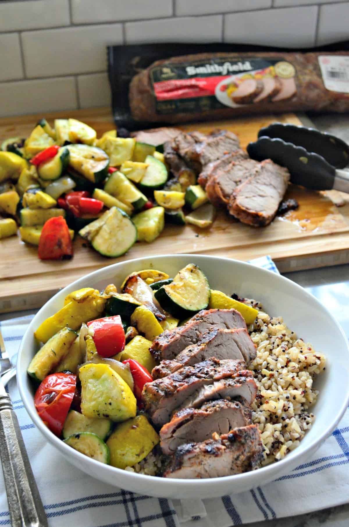 Pork Tenderloin and Brown Rice in a bowl with grilled squash, tomatoes, onions in front of cutting board.