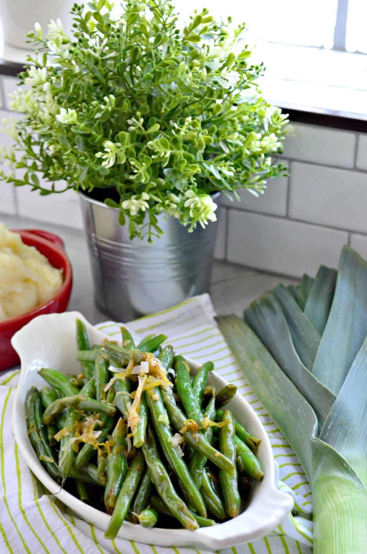 Sauteed Green Beans & Leeks in bowl topped with shredded cheese near bowl of mashed potatoes and fresh leek.