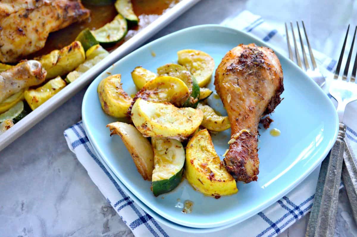 Chicken drumstick with sliced squash and onion on light blue square plate next to forks.
