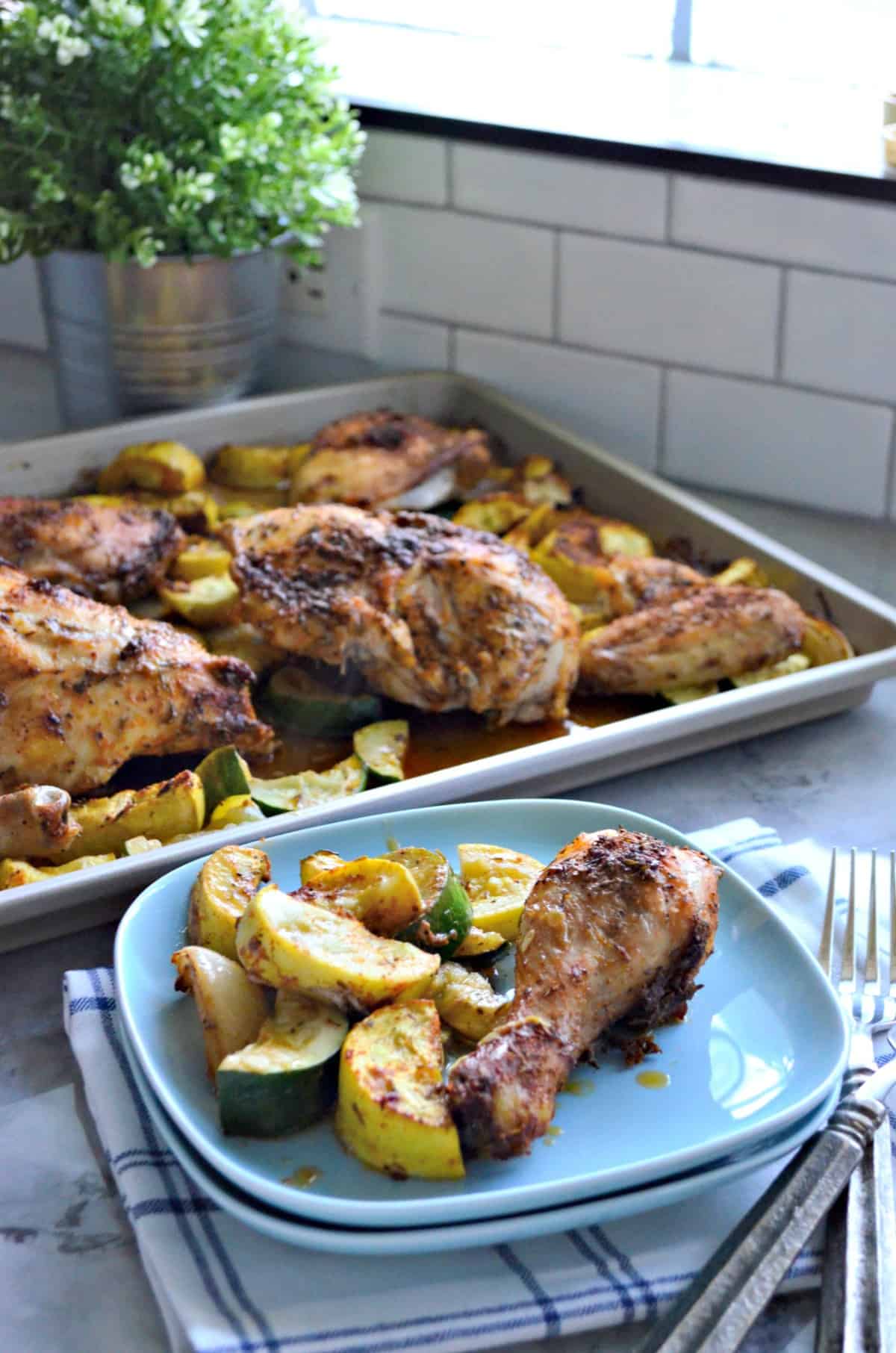 Chicken drumstick with sliced squash and onion on light blue square plate in front of sheet pan.