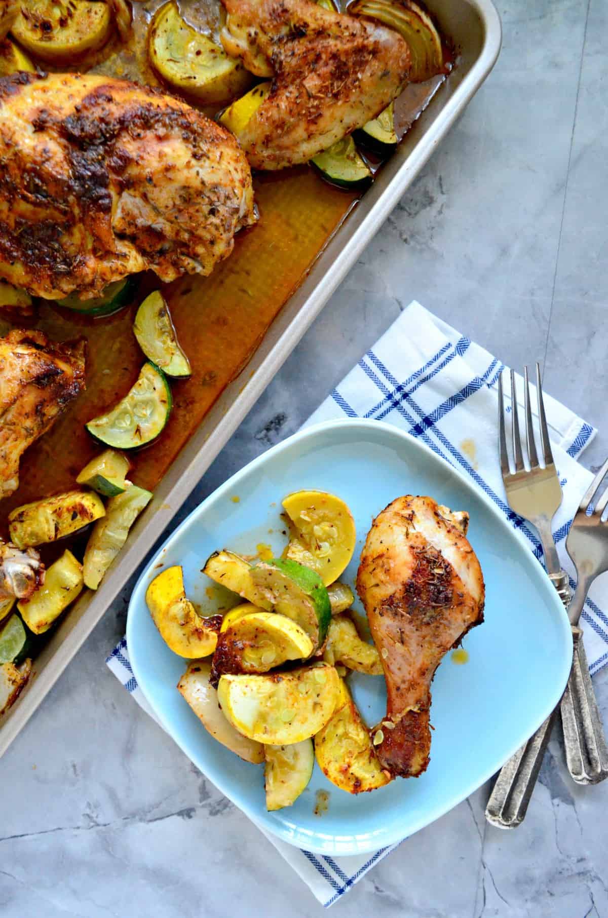 Chicken drumstick with sliced squash and onion on light blue square plate next to sheet pan.