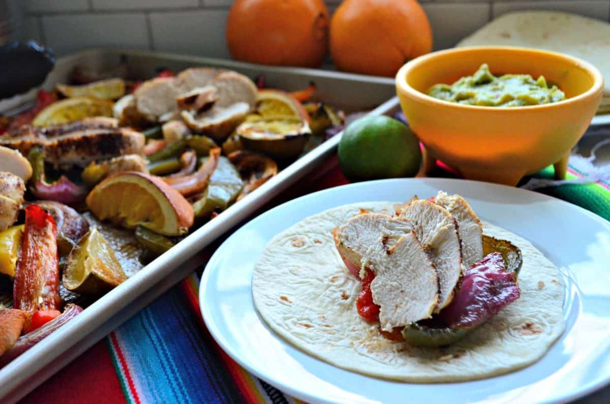 sliced bell peppers, sliced chicken, lemon, and onion on sheet pan next to plated fajita.