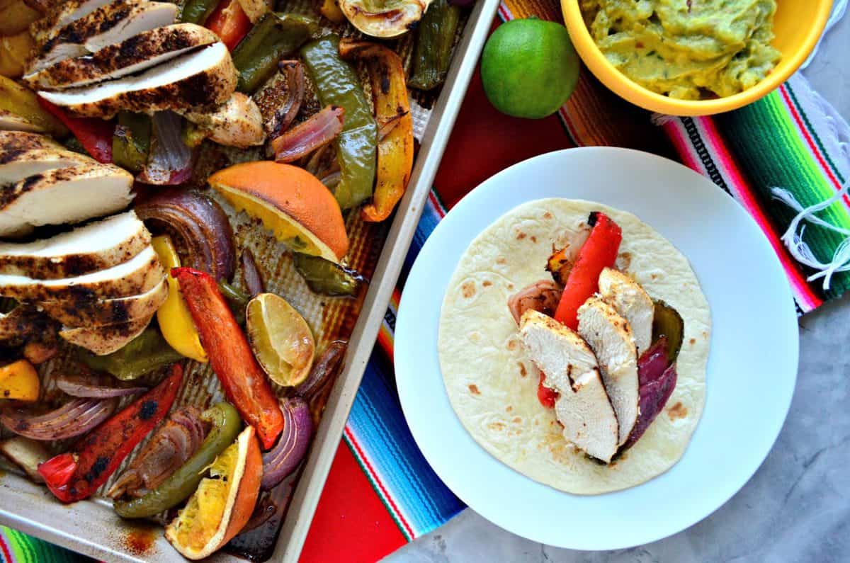 top view of sliced bell peppers, sliced chicken, lemon, and onion on sheet pan next to plated fajita.