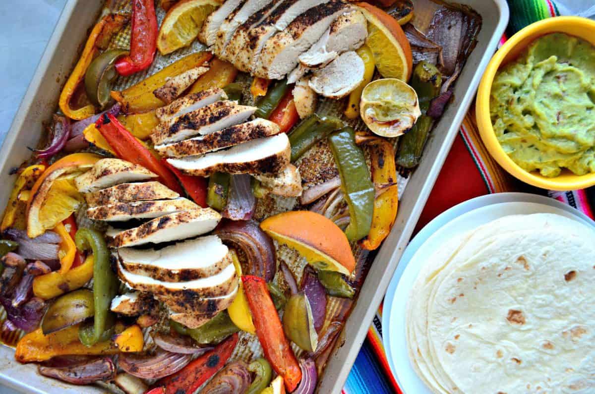 top view of sliced bell peppers, sliced chicken, lemon, and onion on sheet pan next to tortillas.