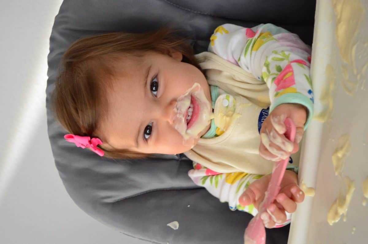 young girl (around 13 months) at high chair smiling with yogurt all over her face with spoon in hand.