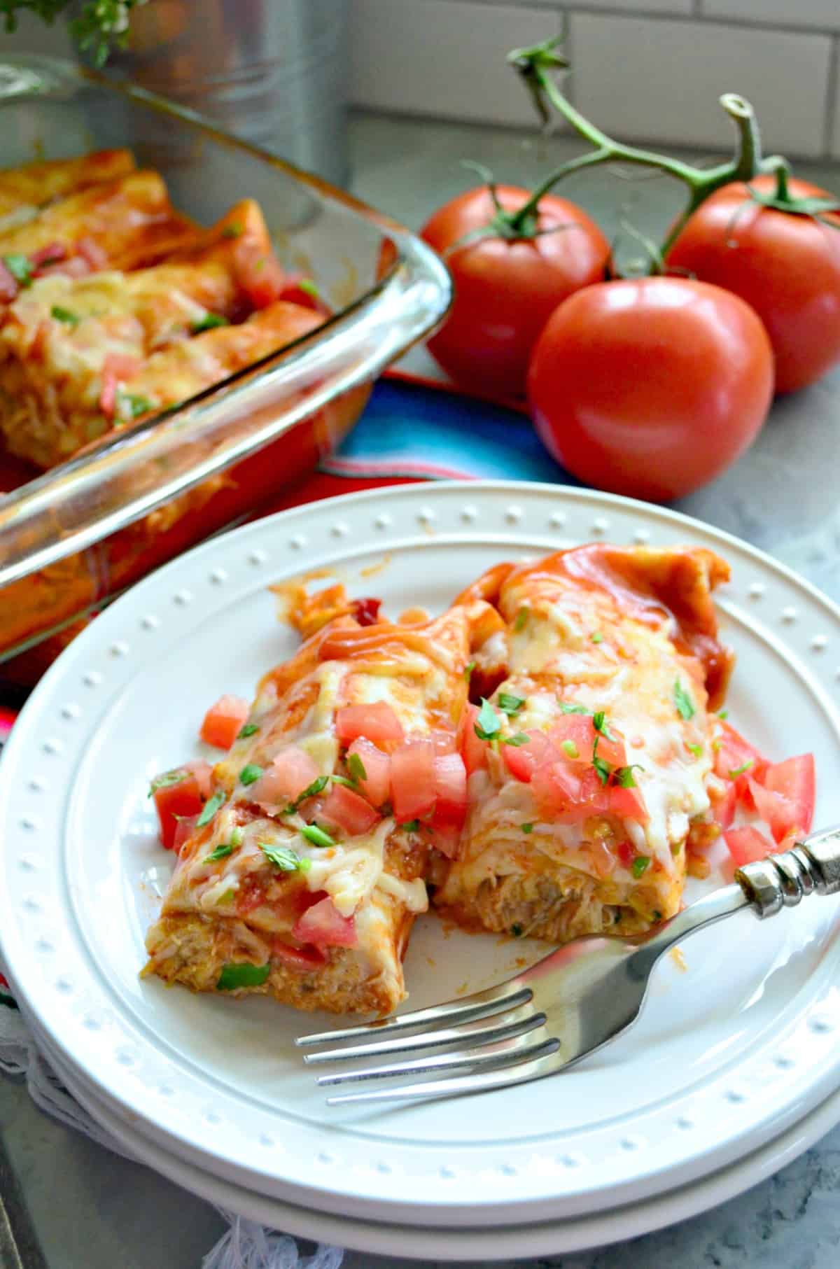 Half of 2 Enchiladas on plate topped with melted cheese, diced tomatoes, and cilantro.