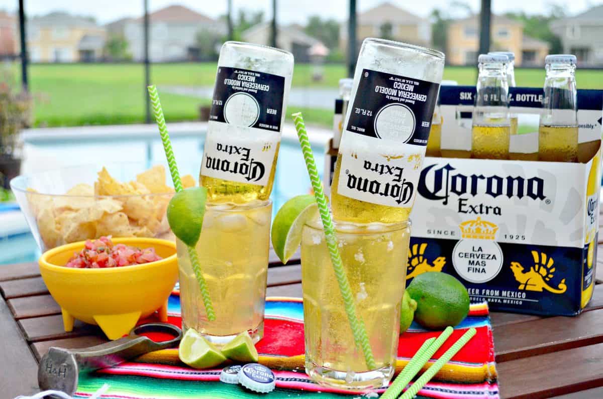 two glasses of light yellow iced liquid with upsidedown corona bottles in them next to chips and salsa.