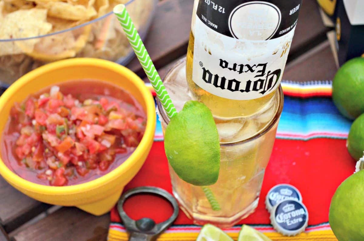 glass of light yellow iced liquid with upsidedown corona bottle in it next to bowl of red salsa and chips.