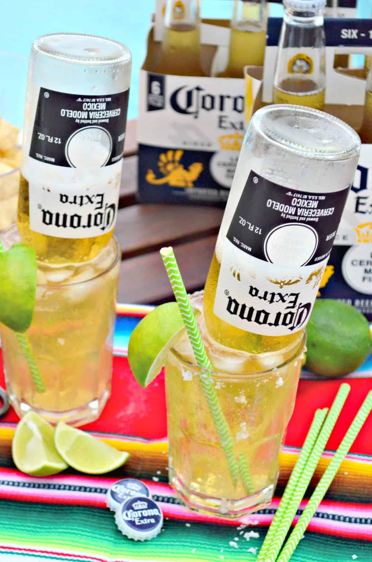 two glasses of light yellow iced liquid with upsidedown corona bottles in them with lime wedges on rim.