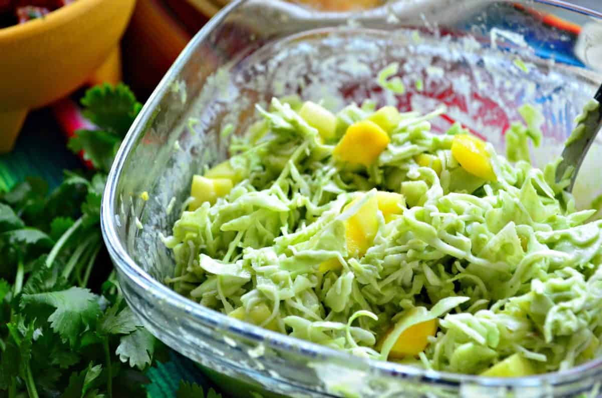 Top view of shredded green bits in glass bowl with cubed mango and green sauce next to cilantro.