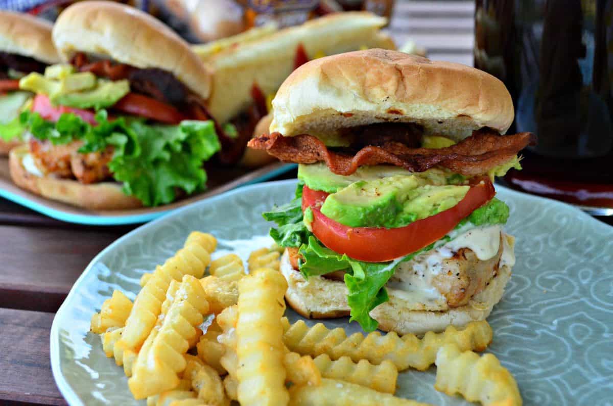 closeup chicken, ranch, lettuce, tomato, bacon, and avocado on a bun with fries on a plate.