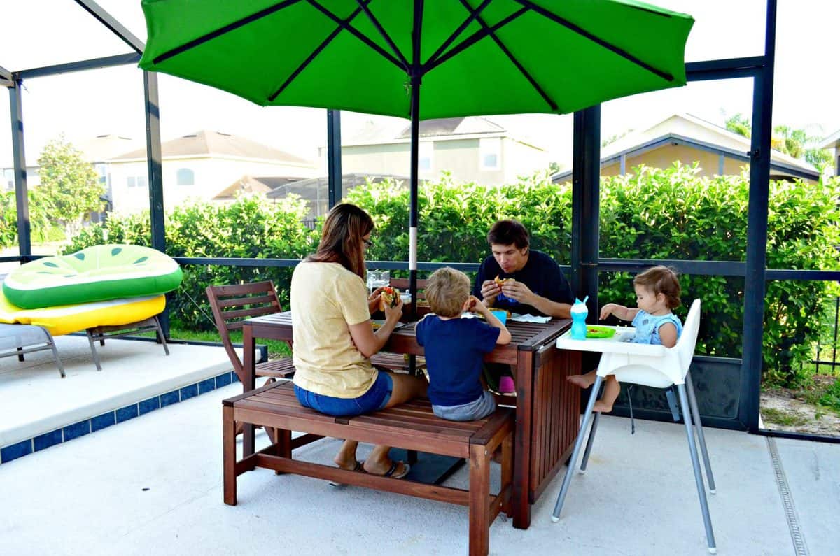 a mom, dad, son, and young daughter enjoying sandwiches at a picnic table with umbrella on backyard pool patio.