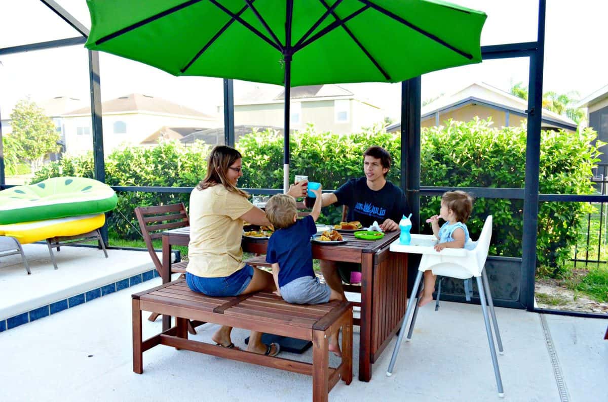 a mom, dad, son, and young daughter cheersing at a picnic table with umbrella as they enjoy lunch.