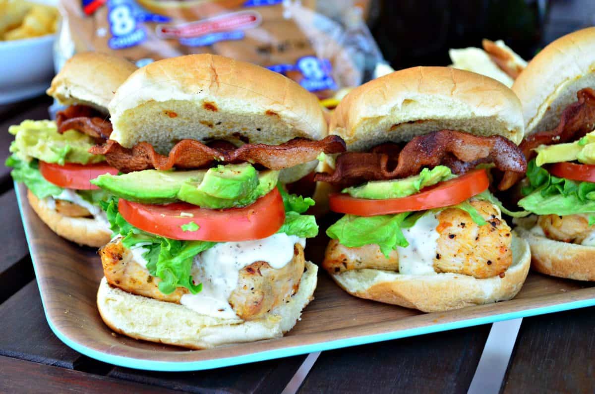 side view of platter of chicken sandwiches on buns with ranch, tomato, lettuce, avocado, and bacon.